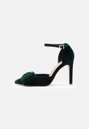 Velvet green high-heeled shoe with a pointed toe, ankle strap, and a floral accent detail at the front. Smooth texture and sleek design.