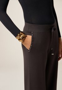 Donkergrijze broeken van zachte stof met zijzakken versierd met metalen studs. Gecombineerd met een aansluitend langsgolftop en een glanzende gouden armband.