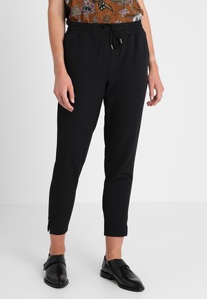Pantalon noir à taille élastique avec poches latérales, coupe slim, longueur cheville, texture de tissu lisse, associé à des chaussures noires perforées.