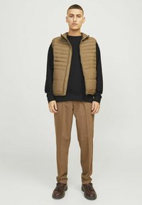 Gilet marrone sopra una felpa nera, abbinato a pantaloni marroni e scarpe scure. Texture lisce, design funzionale e look casual.