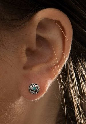 Oreille avec une boucle d'oreille clou florale en argent ornée d'un petit centre turquoise, entourée de détails foncés sur une personne à la peau claire et aux cheveux bruns.