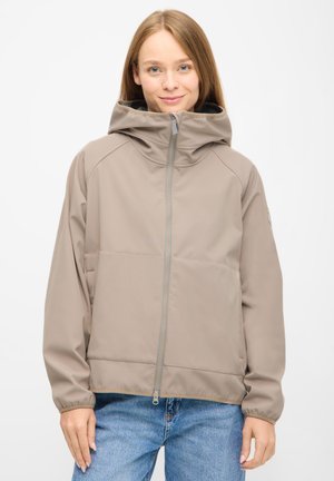 Ung kvinde iført en beige hættejakke, der er lynet op, stående mod en ensfarvet hvid baggrund med hænderne afslappet ned langs siderne.