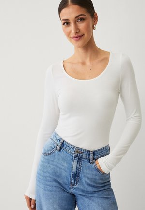 Unge kvinde med mørkt hår iført en hvid langærmet bluse med rund hals og højtaljede lyseblå jeans, hånden i lommen, smilende blidt.