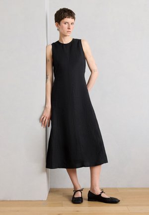 Femme aux cheveux courts, portant une robe midi noire sans manches et des ballerines plates noires à lacets, appuyée d'une main contre un mur gris clair.