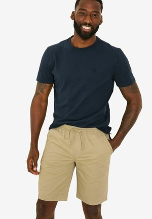 Marineblaue Baumwoll-T-Shirt mit Rundhalsausschnitt und dezentem Logo, kombiniert mit knie-langen beigen Shorts mit elastischem Bund und seitlichen Taschen.