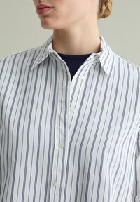 Camicia button-up bianca con righe verticali marine. Presenta un colletto classico, maniche lunghe e chiusura a bottoni. Tessuto dalla texture liscia.