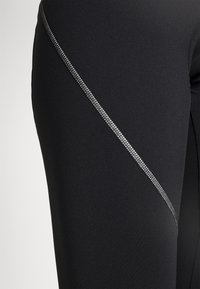 Leggings deportivos negros con una textura suave, que presentan un detalle en costura diagonal reflectante en plata a lo largo del costado.