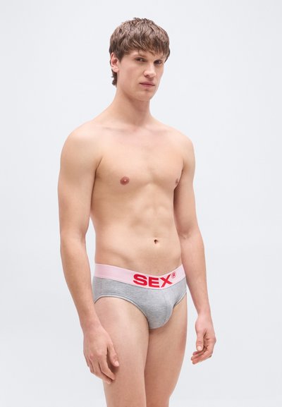 Sive bombažne spodnje hlačke z roza pasom, na katerem je napis "SEX" v rdečih velikih črkah. Modelka je upodobljena v nevtralnem položaju na enobarvnem ozadju.