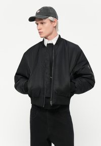 Chaqueta bomber negra con cremallera frontal, puños y dobladillo acanalados; presenta un acabado de satén suave. Combinada con una gorra oscura y una camisa blanca.
