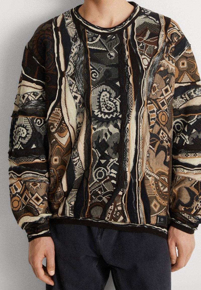 Sweter z dzianiny, łączący odcienie brązu, czerni i kremu, z geometrycznymi i abstrakcyjnymi wzorami. Lekki, z okrągłym dekoltem i ściągaczem na dole.