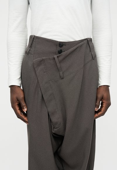 Άνδρας που φοράει γκρι παντελόνια ψηλής μέσης με ασύμμετρο μπροστινό καπάκι και μαύρα κουμπιά, σε συνδυασμό με λευκό πουκάμισο μακρυμάνικο.