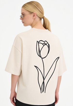Femme blonde avec queue de cheval portant des lunettes de soleil et un t-shirt beige oversize avec un grand dessin linéaire noir de tulipe dans le dos.
