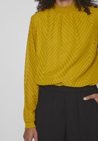 Blouse jaune moutarde avec un motif chevron texturé, des manches longues et un col haut froncé, associée à un pantalon noir à taille élastique.