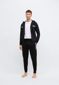 Czarna bluza z kapturem zapinana na zamek, z złotymi paskami i logo, połączona z dopasowanymi czarnymi spodniami dresowymi z bocznymi kieszeniami na zamek. Brak widocznego obuwia.