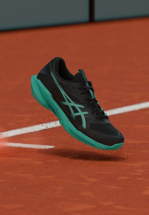 Black and teal athletic shoe lifting off a clay tennis court with red clay particles scattering behind it.