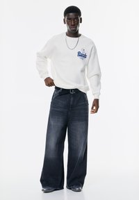 Witte sweatshirt met grafische tekst, ontspannen pasvorm, gecombineerd met donkerblauwe wijde jeans, met een zwarte riem en stijlvolle schoenen.