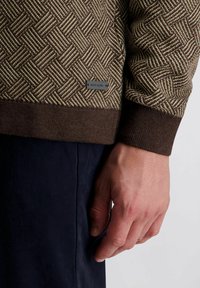 Texturierter brauner Pullover mit einem gewebten geometrischen Muster. Gerippte, dunkelbraune Bündchen und Saum. Kleines Markenlabel an der Seite sichtbar. Teilweise sichtbare Hand.