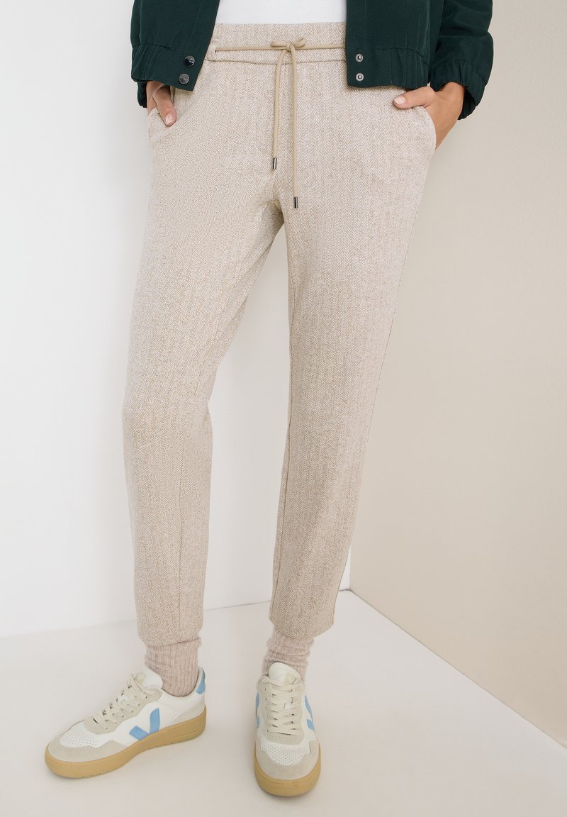 Beige pletené tepláky s strukturovaným vzorem herringbone, elastickými manžetami, stahovacími šňůrkami v pase a předními kapsami. V kombinaci s bílými teniskami s modrými akcenty.