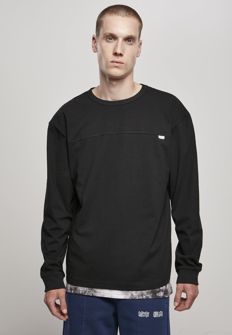 Sort langærmet t-shirt med rund hals og en vandret søm over brystet, parret med blå bukser med hvid tekst.