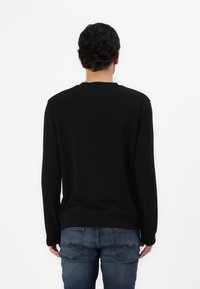 Fekete, hosszú ujjú crewneck pulóver, puha anyagból készült. Egyenes aljú, a háta pedig diszkrét logó részlettel van ellátva. Kék farmernadrággal párosítva.