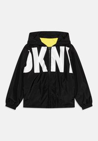 Mustat tuulitakki, jossa on suuri valkoinen "DKNY" -logo, huppu, keltainen vuori, vetoketjukiinnitys, elastiset hihansuut ja kevyt nailonmateriaali.