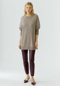 Hellbraune Stricktunika mit langen Ärmeln, gerippten Bündchen und zwei vorderen Taschen. Kombiniert mit burgunderfarbenen Lederleggings und glänzenden, spitzen Schuhen.