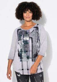 T-shirt gris à manches longues présentant des motifs de texte variés en vert foncé et noir, avec une coupe ample et un col rond.