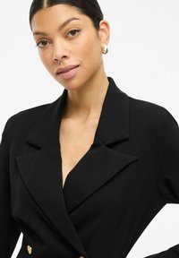 Schwarzer Blazer mit tiefem V-Ausschnitt, großen Revers und doppelten goldenen Knopf-Details. Aus glattem Stoff mit taillierter Passform gefertigt.