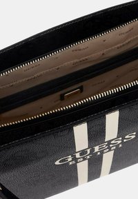 Borsa nera con strisce bianche, con una cerniera aperta che rivela un interno beige con il nome del brand "GUESS" stampato all'interno.