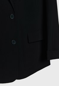 Veste noire cintrée avec deux boutons, une poche à rabat sur le côté droit et des manches retroussées visibles sur un fond uni.