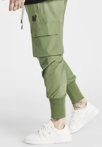 Grüne Cargohosen mit gerippten Bündchen und Fronttaschen, kombiniert mit weißen Turnschuhen. Der Stoff wirkt glatt, mit einem gestickten Logo-Detail.