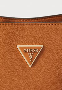 Borsa in pelle marrone con texture, caratterizzata da un logo triangolare in metallo di Guess USA con lettere in rilievo e dettaglio a forma di punto interrogativo.