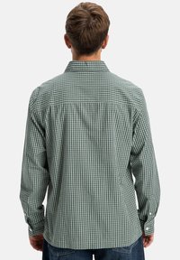 Mann in grün-weißer kariertem Langarmhemd, der mit dem Rücken zur Kamera steht, die Hände an den Seiten, vor einem einfarbigen Hintergrund.