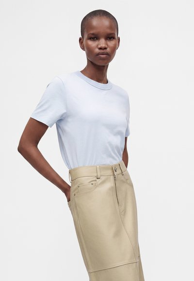Femme aux cheveux courts portant une chemise bleu clair à manches courtes rentrée dans une jupe beige taille haute avec des poches.