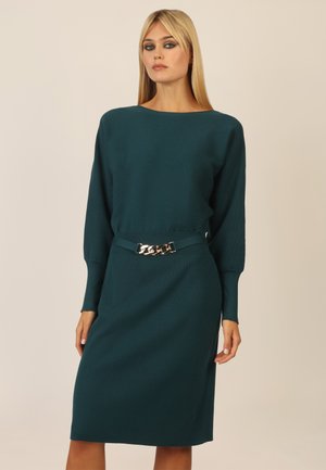 Türkises Strickkleid mit Bootsausschnitt, langen Ärmeln, gerippter Textur und einem goldenen Ketten Gürtel. Figurbetonte Silhouette bis zum Knie.