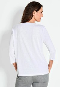 Weißes Langarmshirt mit Rundhalsausschnitt und lockerer Passform, strukturierter Stoff und ein Saum, der unterhalb der Hüften endet, kombiniert mit grauen Hosen.