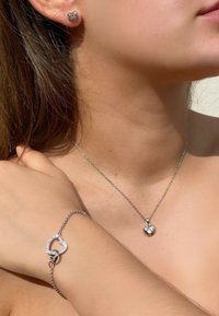 Boucles d'oreilles en argent en forme de cœur, collier pendentif et bracelet. Le collier présente un petit cœur avec une pierre précieuse. Le bracelet a un design de cœurs entrelacés.