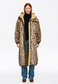 Mujer con cabello rojo que lleva un abrigo largo de piel con estampado de leopardo sobre un suéter de cuello alto negro, jeans azules y zapatos marrones sobre un fondo blanco.