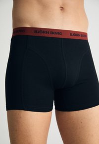 Black boxer briefs made of soft fabric, featuring a burgundy elastic waistband with “BJÖRN BORG” logo. Close-fitting design and smooth texture.