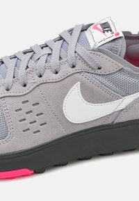 Nike sneakers featuring a gray suede and mesh upper, white swoosh logo, black rubber outsole, and pink accents on the sole.