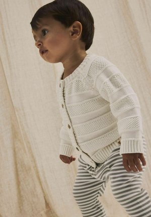 Cardigan bianco lavorato a maglia con strisce testurizzate, dotato di bottoni in legno. Abbinato a leggins a righe grigie e bianche.