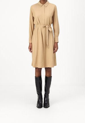 Beige mekko, jossa on vyö, kaulus, pitkät hihat ja sileä rakenne. Yhdistetty mustiin polven yli ulottuviin saappaisiin, joissa on pehmeä, kiiltävä pinta.