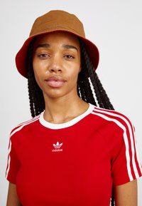 Sarkans pieguļošs T-krekls ar baltām svītrām un Adidas logo, apvienots ar brūnu ķirbju cepuri ar sarkanu oderi, uz vienkārša fona.