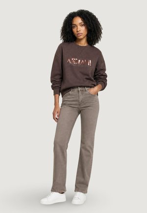 Kvinde med krøllet hår iført mørkebrun Armani sweatshirt, beige jeans med lige ben, hvide sneakers, står med den ene hånd i lommen.