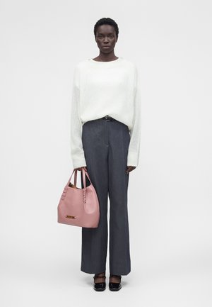 Woman wearing a white sweater and gray wide-leg pants, holding a pink handbag, standing against a plain light background.
