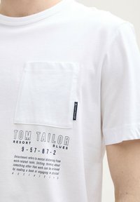 Camiseta de algodón blanca con bolsillo en el pecho izquierdo, escote redondo y diseño de texto impreso en negro. Etiqueta de la marca en la costura lateral.