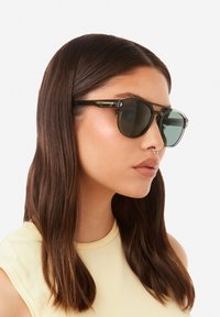 Mustat aurinkolasit, joissa on tummennetut linssit, suorakulmainen muoto ja vihreät yksityiskohdat; malli, jolla on suorat, ruskeat hiukset, käyttää niitä.