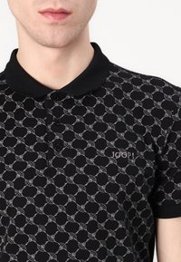 Svart polo-skjorte med et geometrisk mønster i grått, tre-knapp knappestolpe og et brodert logo på venstre bryst. Myk stofftekstur.