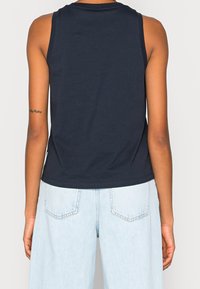 Person som bär en ärmlös marinblå topp och ljusblå denimjeans, visad bakifrån med en liten tatuering på vänster arm.