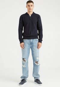 Suéter de punto azul marino con cuello henley y tres botones, combinado con jeans claros desgastados que presentan grandes desgastes. Zapatos grises.